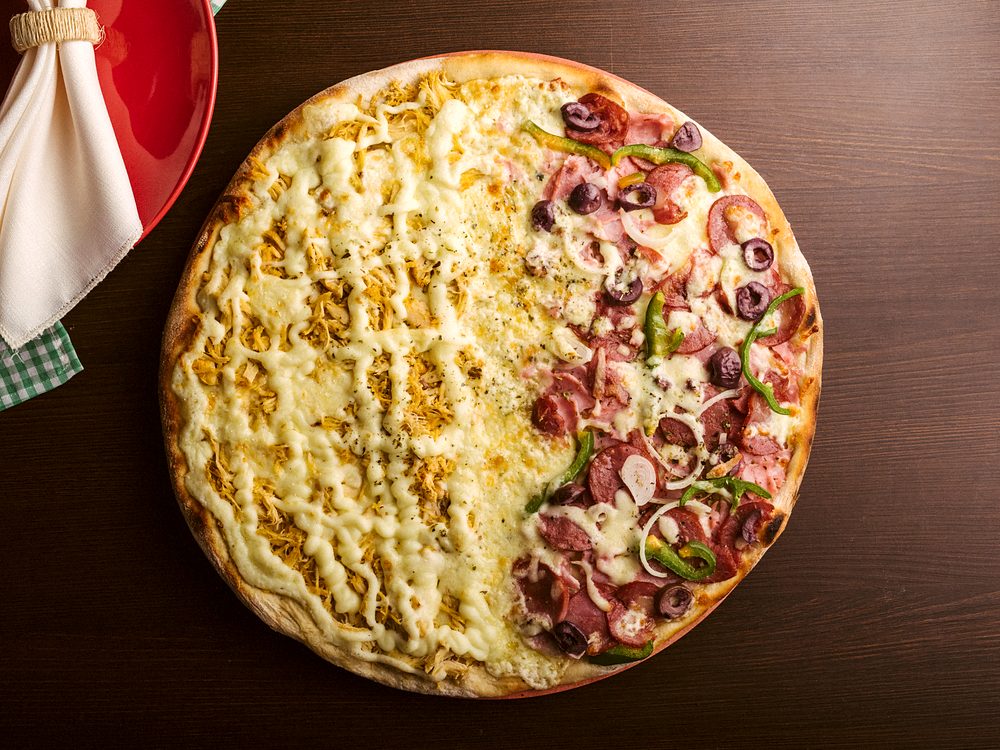 Pizza meio a meio vista de cima, metade com cobertura cremosa e dourada e metade de calabresa com pimentão e azeitona