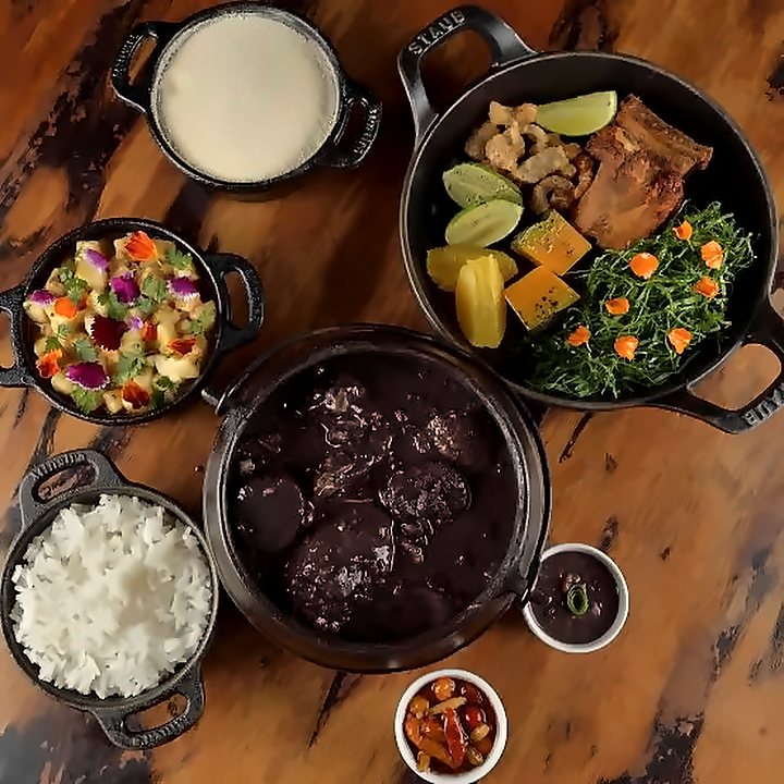 Mesa de feijoada vista de cima, com panelas de feijão, arroz, couve e acompanhamentos